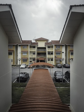 A residential area features a row of yellow apartment buildings with brown tiled roofs. There is a covered walkway leading towards the buildings, surrounded by parked cars and metal lamp posts. The sky is overcast, suggesting cloudy weather.