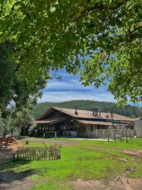 A rustic, wooden building with a sloped roof is surrounded by lush greenery. A clear blue sky is visible above, and there is a wooden fence and some playground equipment on one side. The ground is a mix of grass and dirt.