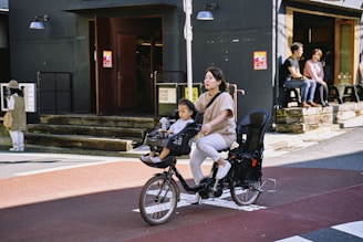A woman rides a bike equipped with seats for children, with a young child seated in front. They are on a street next to a building with dark walls and several people sitting on outdoor benches.