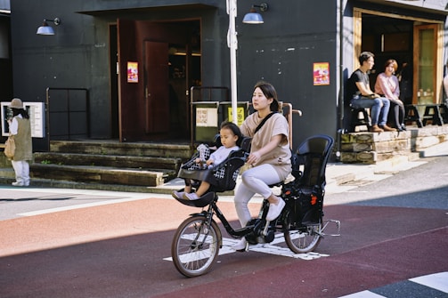 A woman rides a bike equipped with seats for children, with a young child seated in front. They are on a street next to a building with dark walls and several people sitting on outdoor benches.