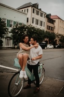 A couple is enjoying a moment together on a city street. The woman is sitting on a teal rental bicycle while embracing the man who stands beside her. They both appear to be in a joyful and affectionate mood. The background features a row of stylish, old-fashioned buildings with large windows and shutters, along with some parked cars and trees lining the street.