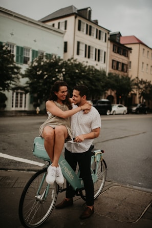 A couple is enjoying a moment together on a city street. The woman is sitting on a teal rental bicycle while embracing the man who stands beside her. They both appear to be in a joyful and affectionate mood. The background features a row of stylish, old-fashioned buildings with large windows and shutters, along with some parked cars and trees lining the street.