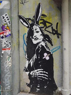 A piece of street art depicting a stylized figure with rabbit ears. The figure appears to be wearing a leather jacket, and their posture is strong and assertive. The background includes various graffiti and stickers, contributing to an urban and edgy atmosphere.