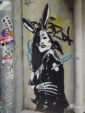A piece of street art depicting a stylized figure with rabbit ears. The figure appears to be wearing a leather jacket, and their posture is strong and assertive. The background includes various graffiti and stickers, contributing to an urban and edgy atmosphere.