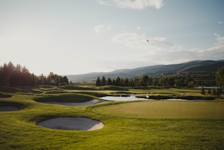 A scenic golf course with well-maintained greens and sand bunkers surrounded by lush greenery. The landscape includes distant hills and a serene sky with scattered clouds. A bird is soaring above, adding to the peaceful ambiance.