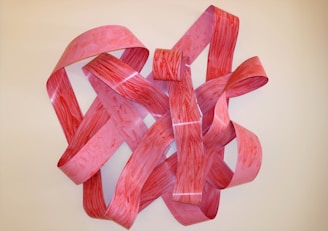 A complex three-dimensional sculpture composed of intertwined, wavy strips of material with a marbled texture. The colors range from pink to red, creating a visually striking effect against a neutral background.