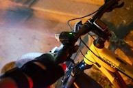 A cyclist wearing gloves grips the handlebars of a Cannondale bicycle. The bike is equipped with a digital display and a small light, indicating night or low-light conditions. The frame color is yellow, and the surroundings appear to be an urban setting with a sidewalk or street.