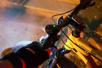 A cyclist wearing gloves grips the handlebars of a Cannondale bicycle. The bike is equipped with a digital display and a small light, indicating night or low-light conditions. The frame color is yellow, and the surroundings appear to be an urban setting with a sidewalk or street.