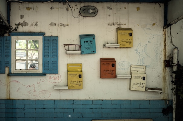 A vintage-style interior wall features multiple colorful mailboxes mounted in an artistic arrangement. A small window with blue shutters is on the left, showing a glimpse of the outdoors. The wall is adorned with whimsical drawings of cats, contributing to a playful atmosphere. Some areas of the wall have peeling paint and visible wear, adding an aged charm to the scene.