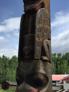 A tall, intricately carved wooden totem pole stands against a backdrop of greenery and a partly cloudy sky. The totem pole features detailed animal and abstract designs, showcasing traditional craftsmanship. In the background, there are trees and a building with a red roof.
