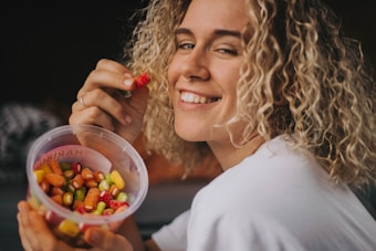 A person with curly blonde hair smiles while holding a container filled with colorful gummy candies. The relaxed atmosphere and casual clothing suggest a lighthearted and joyful moment.