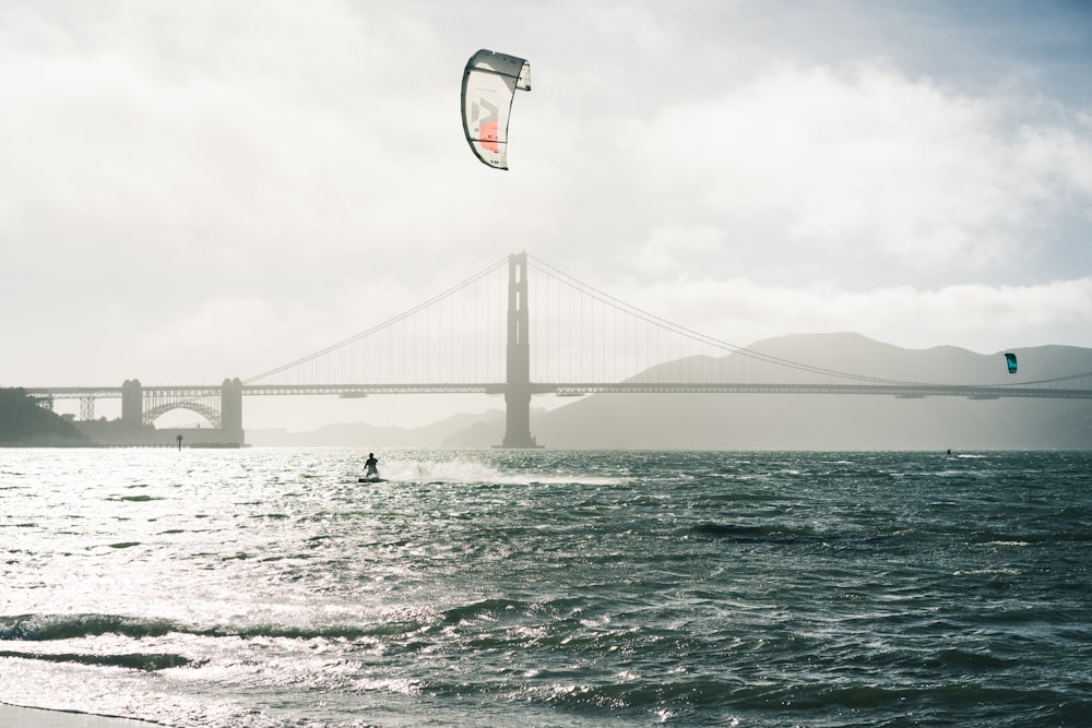 person surfing on sea near bridge under cloudy sky during daytime