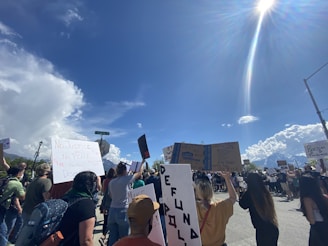 A protest with a diverse crowd holding various signs and banners under a clear blue sky. The signs include messages related to justice and equality. The atmosphere is one of activism and social movement.