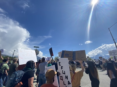 A protest with a diverse crowd holding various signs and banners under a clear blue sky. The signs include messages related to justice and equality. The atmosphere is one of activism and social movement.