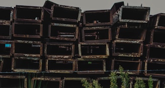 A stack of rusted, rectangular metal beams is arranged in a disorderly manner. The metal surfaces display a coarse, weathered texture with a brownish-red patina. In the foreground, some green plants are visible, adding a touch of nature to the industrial scene.