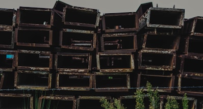 A stack of rusted, rectangular metal beams is arranged in a disorderly manner. The metal surfaces display a coarse, weathered texture with a brownish-red patina. In the foreground, some green plants are visible, adding a touch of nature to the industrial scene.