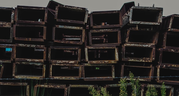 A stack of rusted, rectangular metal beams is arranged in a disorderly manner. The metal surfaces display a coarse, weathered texture with a brownish-red patina. In the foreground, some green plants are visible, adding a touch of nature to the industrial scene.