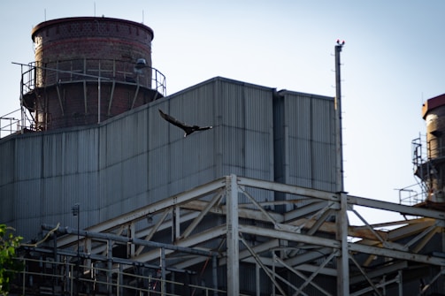 An industrial building with large, metal structures and a brick chimney is seen with a bird flying across the sky.