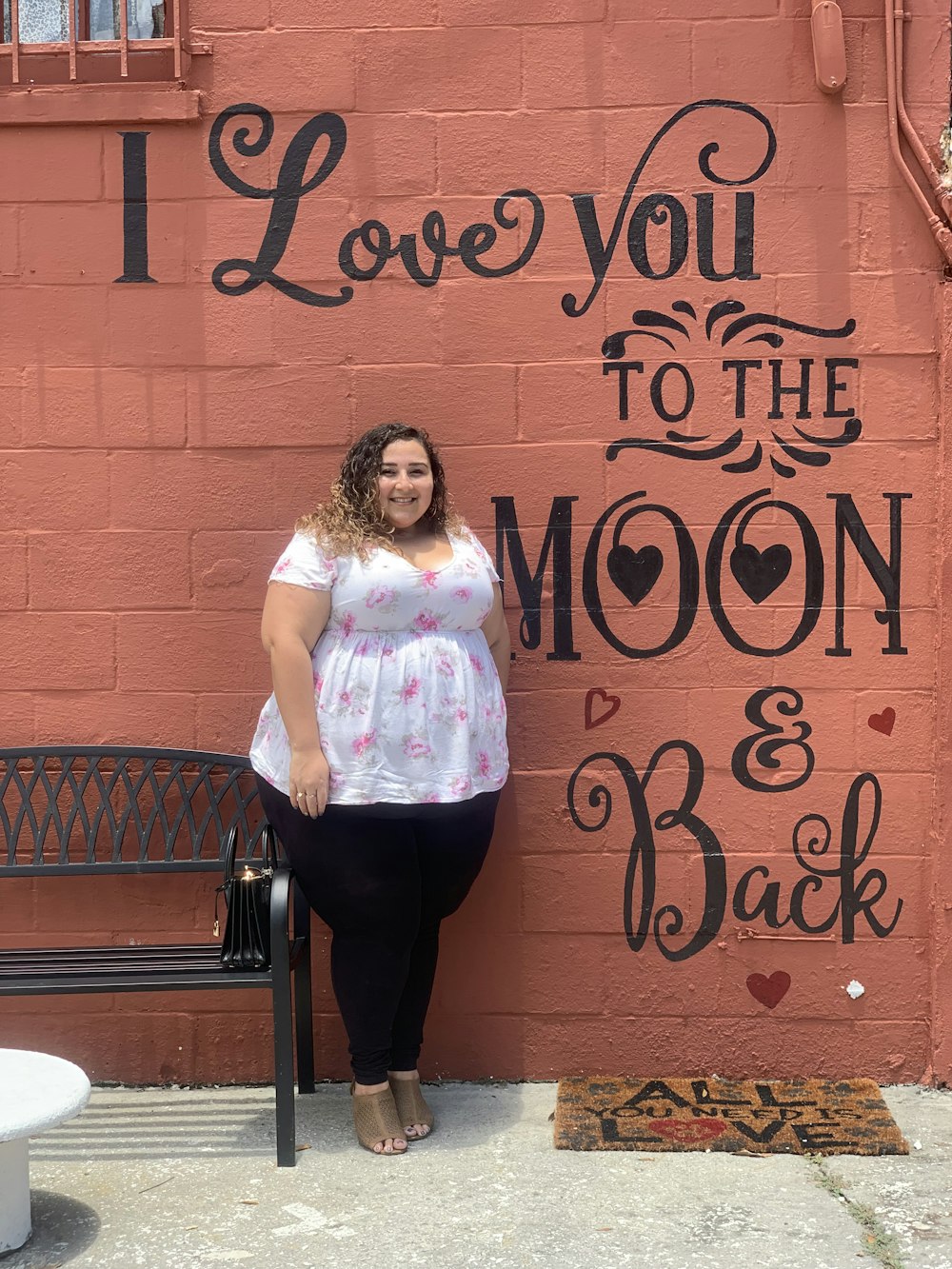 woman in white and pink floral shirt and black pants standing beside brown wooden wall