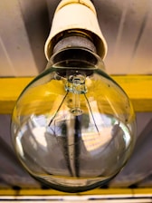 A close-up view of a clear incandescent light bulb mounted on a ceiling. The metal base and the glass bulb are prominently visible, with a slightly reflective surface capturing light and shadows. The internal filament structure of the bulb is apparent, and the fixture appears to be attached to a yellow support structure under a metallic ceiling.