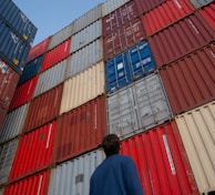 man in black jacket standing in front of red and blue intermodal containers