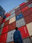 man in black jacket standing in front of red and blue intermodal containers