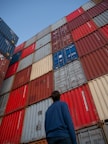 man in black jacket standing in front of red and blue intermodal containers