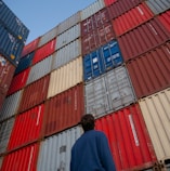 man in black jacket standing in front of red and blue intermodal containers