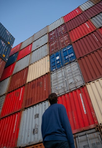 man in black jacket standing in front of red and blue intermodal containers