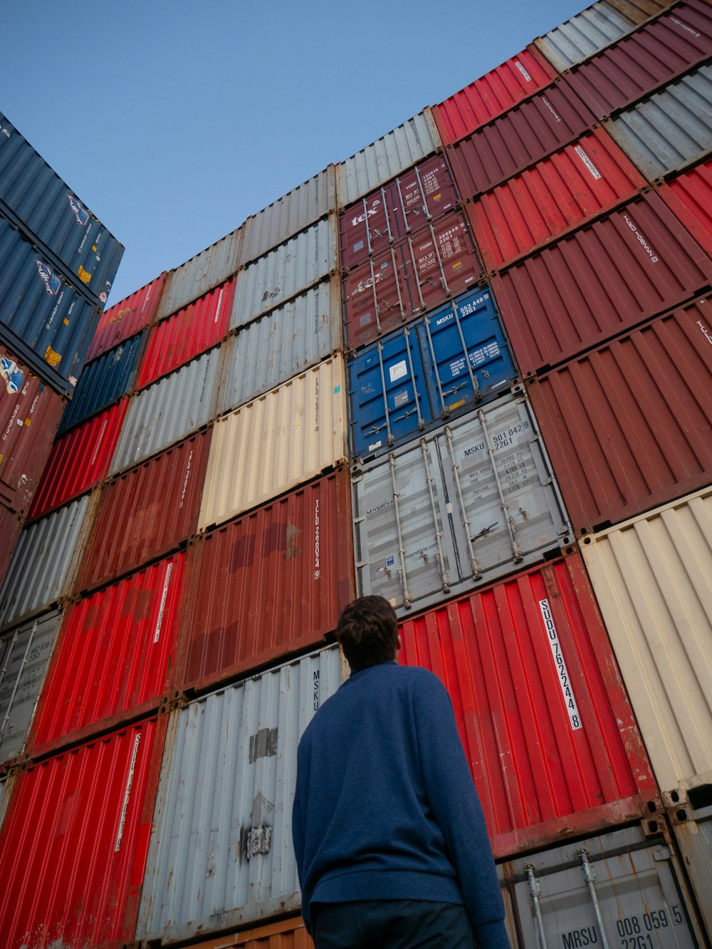 man in black jacket standing in front of red and blue intermodal containers