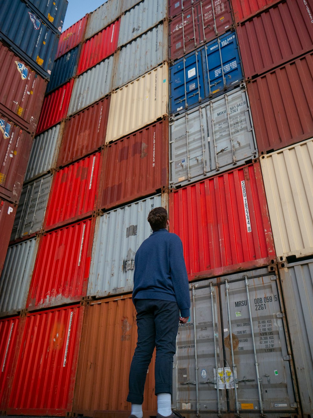 man in blue long sleeve shirt standing in front of red and blue intermodal containers