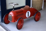 A vintage-style red toy car designed for children, featuring a number one on its side. The car has a glossy finish, black wheels, a chrome steering wheel, and brown leather-like seat. It is positioned on a light-colored floor near a woven basket.