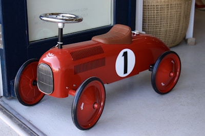 A vintage-style red toy car designed for children, featuring a number one on its side. The car has a glossy finish, black wheels, a chrome steering wheel, and brown leather-like seat. It is positioned on a light-colored floor near a woven basket.