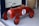 A vintage-style red toy car designed for children, featuring a number one on its side. The car has a glossy finish, black wheels, a chrome steering wheel, and brown leather-like seat. It is positioned on a light-colored floor near a woven basket.