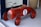 A vintage-style red toy car designed for children, featuring a number one on its side. The car has a glossy finish, black wheels, a chrome steering wheel, and brown leather-like seat. It is positioned on a light-colored floor near a woven basket.