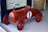 A vintage-style red toy car designed for children, featuring a number one on its side. The car has a glossy finish, black wheels, a chrome steering wheel, and brown leather-like seat. It is positioned on a light-colored floor near a woven basket.
