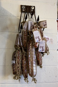 Several cured sausages hang on a wall display, wrapped in netting and secured with tags. The sausages are labeled 'Chorizo' and appear to be part of a specialty meat selection. They are suspended by black hooks against a light-colored brick wall.