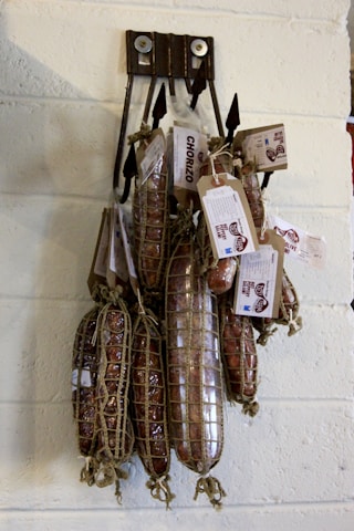 Several cured sausages hang on a wall display, wrapped in netting and secured with tags. The sausages are labeled 'Chorizo' and appear to be part of a specialty meat selection. They are suspended by black hooks against a light-colored brick wall.