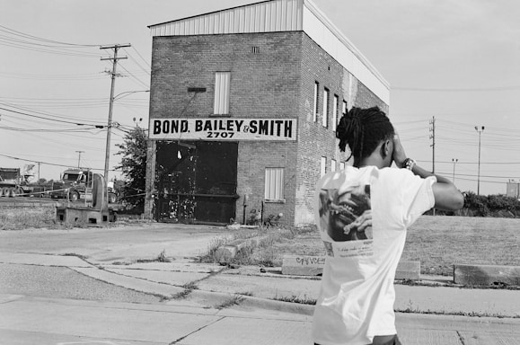 A man is standing on a cracked concrete surface wearing a t-shirt, looking towards a large brick industrial building with a sign reading 'Bond, Bailey & Smith 2707'. There are power lines and another large vehicle visible in the background, and the overall setting appears to be urban and slightly rundown.