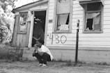 A person crouches in front of an old, dilapidated house with broken windows. The house has peeling paint and a number, 430, spray-painted on the siding. The area is overgrown with grass and plants, suggesting neglect.