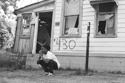 A person crouches in front of an old, dilapidated house with broken windows. The house has peeling paint and a number, 430, spray-painted on the siding. The area is overgrown with grass and plants, suggesting neglect.