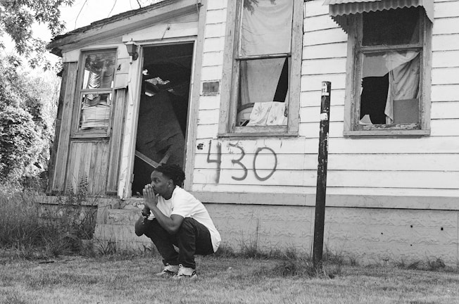 A person crouches in front of an old, dilapidated house with broken windows. The house has peeling paint and a number, 430, spray-painted on the siding. The area is overgrown with grass and plants, suggesting neglect.