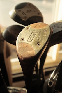 Several vintage golf clubs with wooden heads are positioned close together, showcasing their aged textures and metallic components. The central club features identifiable markings and screws, with sunlight casting a warm glow over the scene.