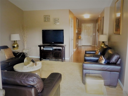 A cozy living room features dark leather sofas and armchairs adorned with shiny gold pillows. A flat-screen TV sits on a wooden stand against a beige wall, surrounded by decorative accents like vases and lamps. A round coffee table holds a small vase and a few decorative items. The space is warmly lit, with a view of the hallway leading to the front door.