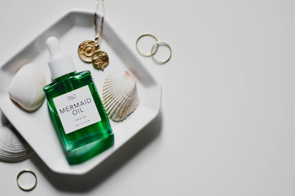 A green bottle labeled 'Mermaid Oil Face Oil' rests on a white geometric dish. Next to the bottle are seashells and gold-toned jewelry, including a pendant and rings. The setting is minimalist with a clean, white background.