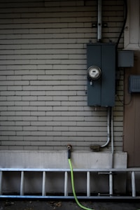 A brick wall painted in a light neutral color has an electrical meter and panel installed on it. A metal ladder is leaning horizontally at the bottom, and a green garden hose is attached to an exterior faucet.