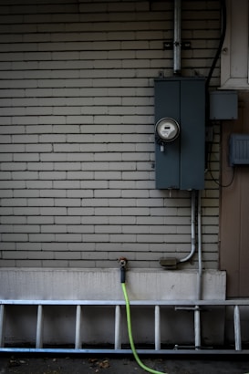 A brick wall painted in a light neutral color has an electrical meter and panel installed on it. A metal ladder is leaning horizontally at the bottom, and a green garden hose is attached to an exterior faucet.