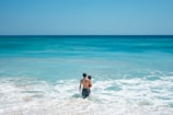 A man, shirtless and wearing dark shorts, is holding a child and standing in the shallow waves of a beach. The ocean stretches out into the horizon, with varying shades of blue water under a clear blue sky. The waves are gently crashing around their legs.