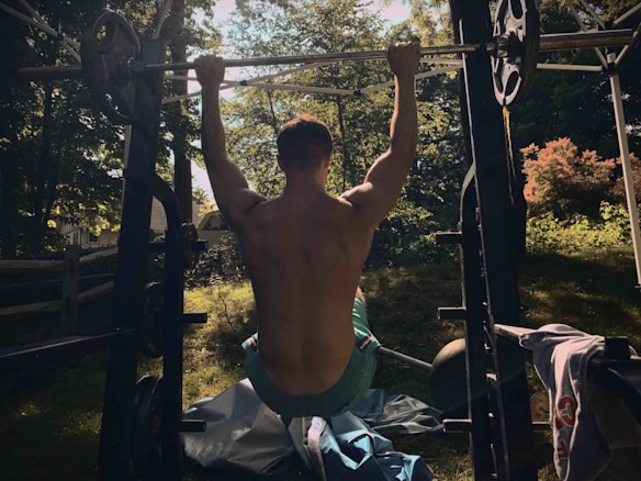 A shirtless person is performing a pull-up on an outdoor gym setup, surrounded by trees and greenery. The lighting creates shadows on the person's back, enhancing the muscular definition. In the background, sunlight filters through the leaves, casting dappled patterns on the ground.