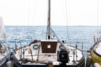 A sailboat is docked at a marina, surrounded by calm blue waters under a bright sky. The boat is equipped with various nautical instruments and ropes, with a wooden cabin door and a number of fenders attached to its sides.